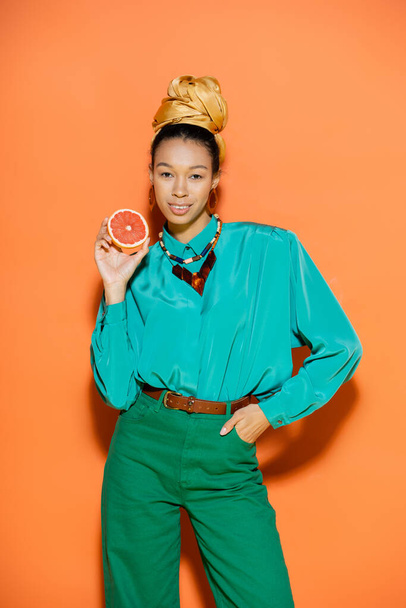 Smiling african american model in summer outfit holding grapefruit on orange background  - Foto, afbeelding