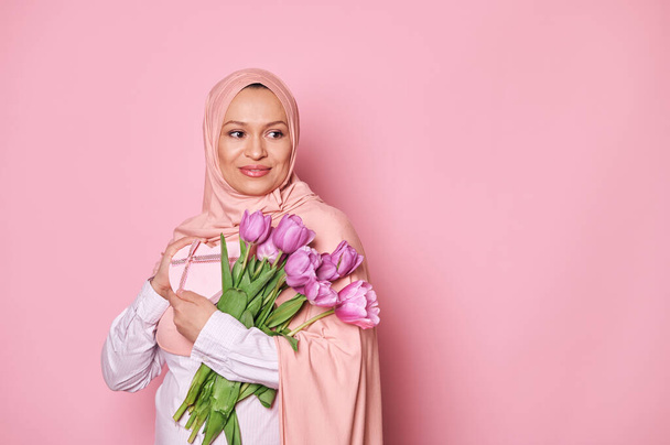 Wunderschöne Muslimin im rosafarbenen Hijab, lächelnd zur Seite blickend, einen Strauß mit lila Tulpen und einer Geschenkschachtel in der Hand, isoliert auf rosa Hintergrund. Internationaler Frauentag und Muttertag. Freier Anzeigenplatz - Foto, Bild
