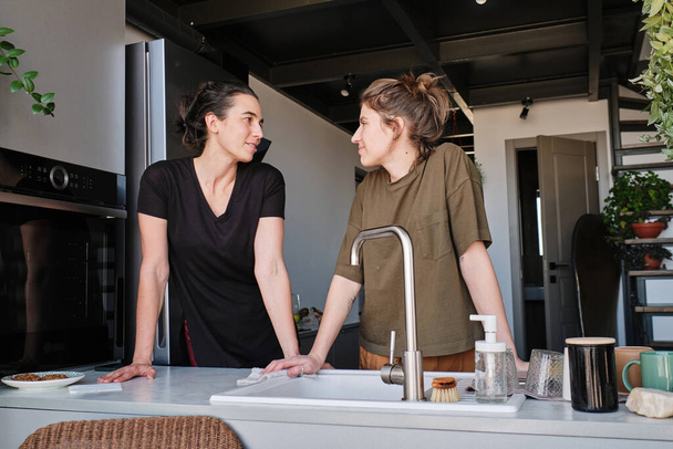Young lesbian couple looking at each other while standing at table with sink during household in kitchen - Photo, Image