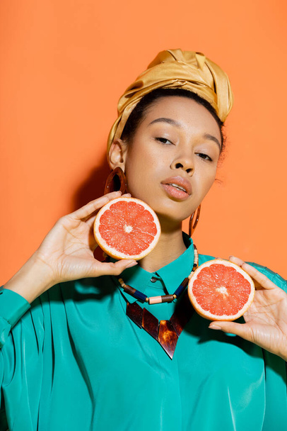 Portrait of stylish african american model with headscarf holding cut grapefruit on orange background  - Foto, afbeelding