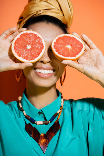 positive and stylish african american model covering face with grapefruit on orange background  - Fotografie, Obrázek