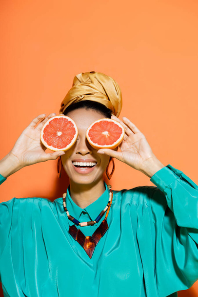 Positive african american woman in summer outfit covering face with fresh grapefruit on orange background  - Foto, Imagem