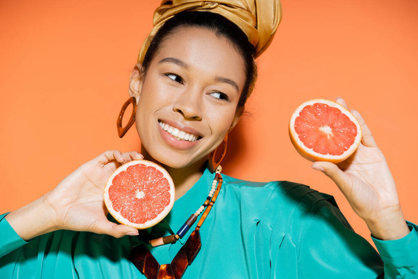 Portrait of positive african american woman in blouse holding cut grapefruit on orange background  - Foto, immagini
