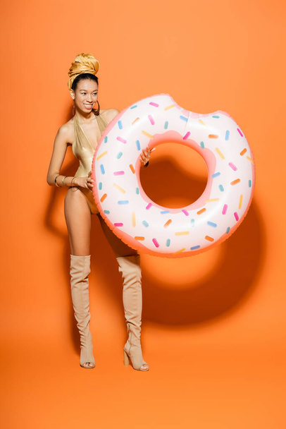 Cheerful and stylish african amercan woman in swimsuit holding pool ring on orange background  - 写真・画像