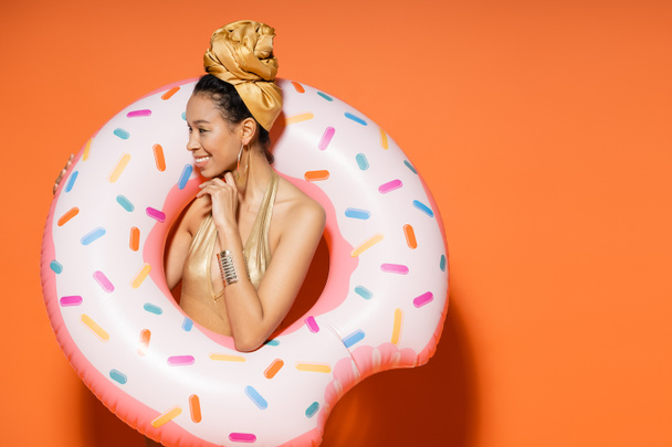 Smiling african american woman in golden headscarf holding pool ring on orange background  - Foto, Imagem