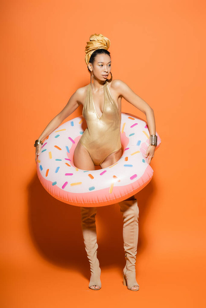 Longitud completa del elegante modelo afroamericano en traje de baño con anillo de piscina sobre fondo naranja  - Foto, imagen