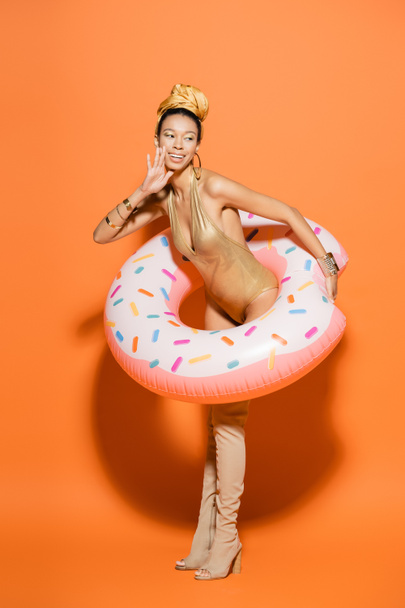 Modelo afroamericano positivo en botas de rodilla y traje de baño con anillo de piscina sobre fondo naranja  - Foto, Imagen