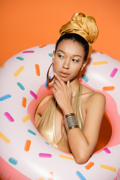 Portrait of pensive african american woman in headscarf posing with pool ring on orange background  - Fotografie, Obrázek