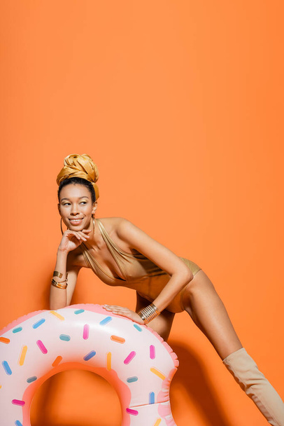 Smiling african american woman in swimsuit posing near pool ring on orange background  - Foto, imagen