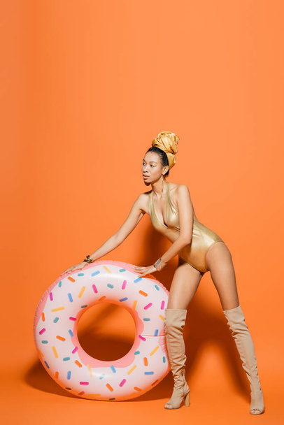 Full length of stylish african american model in golden swimsuit standing near pool ring on orange background  - Photo, Image