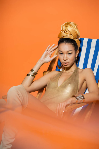 Fashionable african american model in in golden swimsuit sitting on deck chair on orange background  - 写真・画像