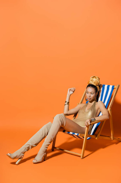 Full length of stylish african american woman in swimsuit and headscarf sitting on deck chair on orange background  - Fotografie, Obrázek