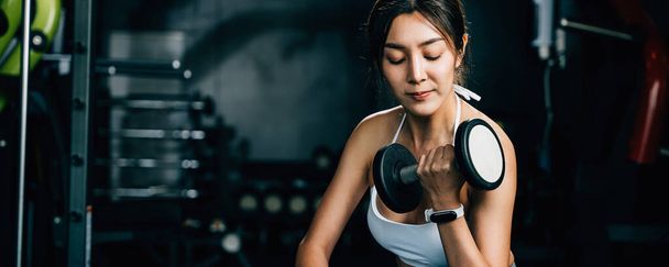 A fitness enthusiast sitting on a gym bench and lifting dumbbells to improve her strength and power for effective weightlifting training, fitness GYM dark background - Fotoğraf, Görsel