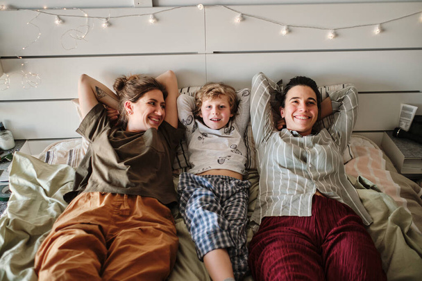 Retrato de familia lesbiana feliz con hijo acostado en la cama y disfrutando del tiempo juntos - Foto, Imagen
