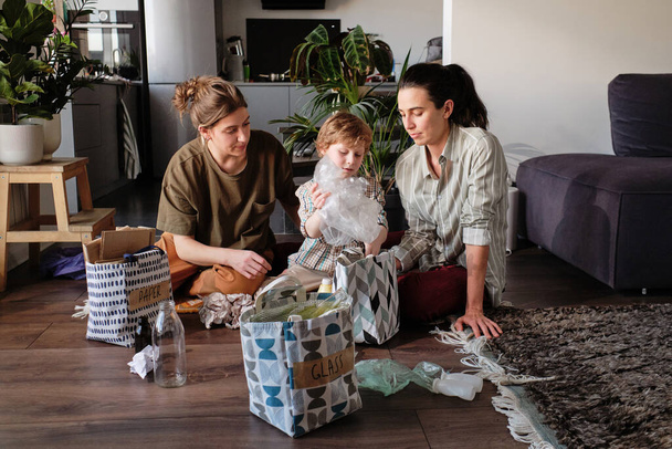 Familia lesbiana con niño pequeño sentado en el suelo en la habitación y la clasificación de la basura en diferentes bolsas - Foto, imagen