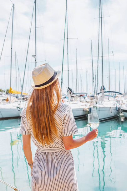 Ξανθιά γυναίκα με καπέλο στέκεται με την πλάτη της στη θάλασσα, γιοτ, κτίρια και βάρκες φόντο. Λευκό ποτήρι κρασιού στο χέρι. Διακοπές στην Ευρώπη. Ωραία, Γαλλική Ριβιέρα. Ταξιδιωτική φωτογραφία. - Φωτογραφία, εικόνα
