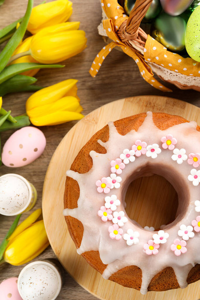 Delicious Easter cake decorated with sprinkles near painted eggs and tulips on wooden table, flat lay - Photo, Image
