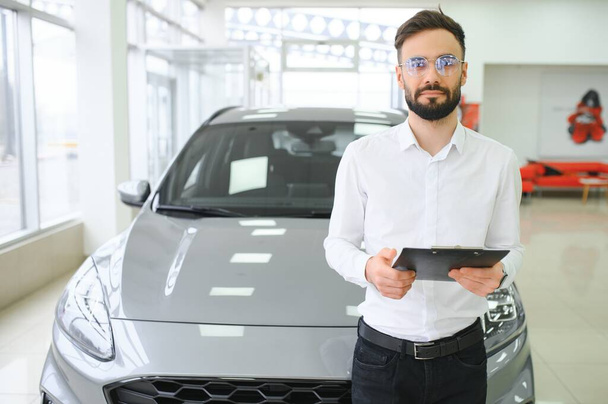 Closeup shot of successful confident smiling caucasian male shop assistant holding clipboard in formal clothes at automobile car dealer shop. - Фото, зображення