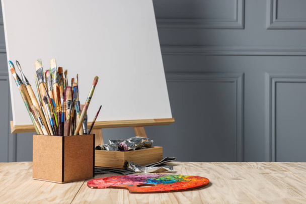 Easel with blank canvas and different art supplies on wooden table near grey wall. Space for text - Foto, Bild