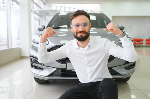 Glücklicher junger Kerl, der ein neues Luxusauto checkt und im Autohaus ein Auto kauft. Porträt eines fröhlichen Millennial-Kaukasiers, der das Auto im Showroom untersucht. - Foto, Bild