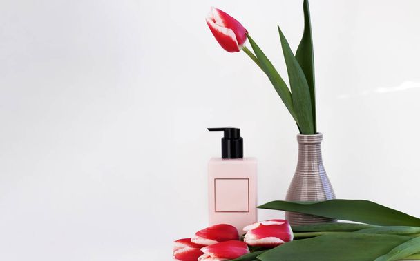 pink bottle with frame in middle for text empty space with tulips flowers isolated on pink or gray background.cosmetic bottle body care cream lotion.flowers petals textile red dark material.laptop - Photo, Image