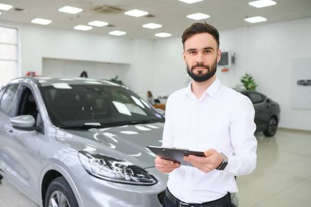 Closeup shot of successful confident smiling caucasian male shop assistant holding clipboard in formal clothes at automobile car dealer shop. - Foto, afbeelding