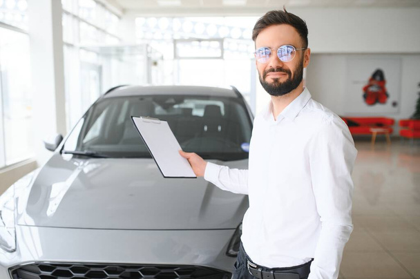 Confident young caucasian businessman shop assistant standing in car automobile dealer shop. - Foto, imagen