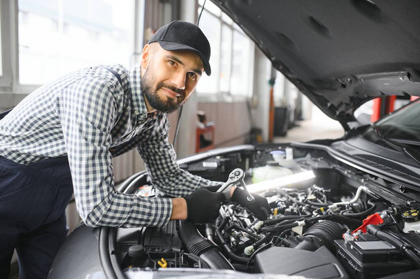 Professional Mechanic is Working on a Car in a Car Service. Repairman in Safety Glasses is Fixing the Engine on a Vehicle. Specialist Unscrews Bolts with a Ratchet. Modern Clean Workshop. - Zdjęcie, obraz