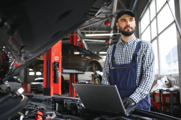 A bearded auto-mechanic in working uniform is standing next to a broken car and using the special laptop for car maintenance. An auto-mechanic in workshop repairs car - Fotoğraf, Görsel