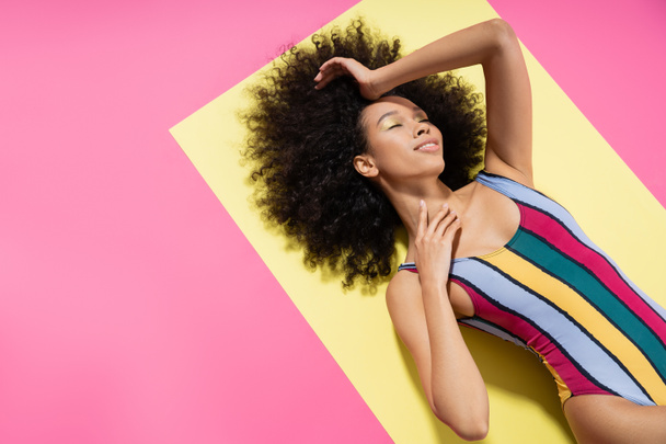 vista dall'alto del modello americano africano soddisfatto in costume da bagno colorato a righe sdraiato su sfondo giallo e rosa  - Foto, immagini