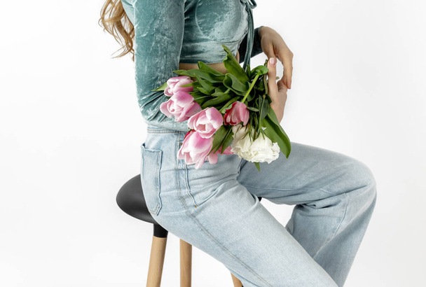 schöne zarte Frau hält Tulpen Strauß in den Armen sitzt auf Stuhl mit Blumen durch Beine in weißem Rock.lange blonde Haare weiblich auf Hochstuhl im Studio isoliert in blauen Jeans Rückansicht - Foto, Bild