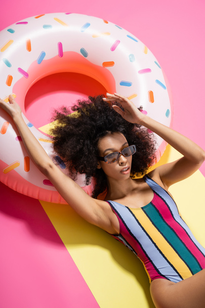 top view of charming african american model in striped swimwear and sunglasses getting tan near inflatable ring on yellow and pink  - Fotografie, Obrázek