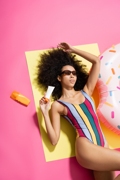 top view of african american woman in striped swimwear and trendy sunglasses holding sunscreen near inflatable ring on pink - Φωτογραφία, εικόνα