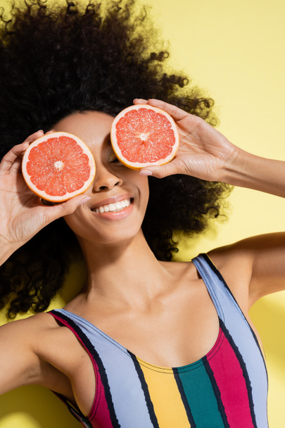top view of happy african american woman in colorful swimsuit covering eyes with grapefruit halves on yellow background  - Foto, immagini