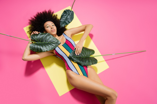 top view of curly african american woman in colorful swimsuit getting tan near green palm leaves on yellow and pink  - Foto, immagini