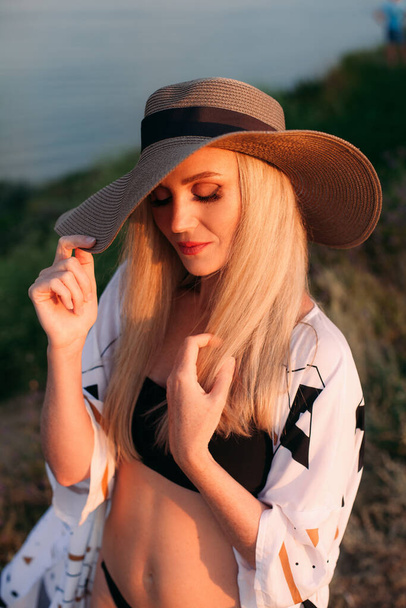 Portrait of a beautiful blonde girl with long hair in a black swimsuit, a light pareo and a hat, near the ocean, at sunset. - Фото, зображення
