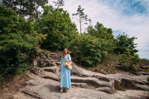 Smukła, młoda dziewczyna w niebieskiej sukience stoi na kamieniach w górach, w słoneczny dzień. W rękach trzyma różowy kapelusz i beżową torebkę.. - Zdjęcie, obraz
