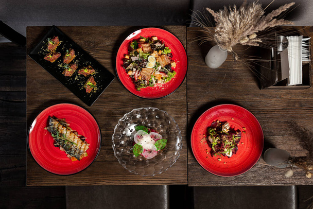 Mittagessen mit ukrainischen Gerichten. Barschtartar, Kalbstartar, gegrillte Makrele, Caesar-Salat und Rote-Bete-Salat liegen auf Keramiktellern auf dunklem, hölzernem Hintergrund. - Foto, Bild