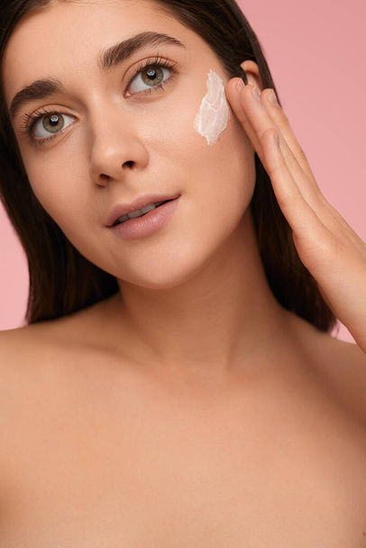Crop young female with bare shoulders and applying moisturizing cream on face, while looking at camera on pink background - Photo, image