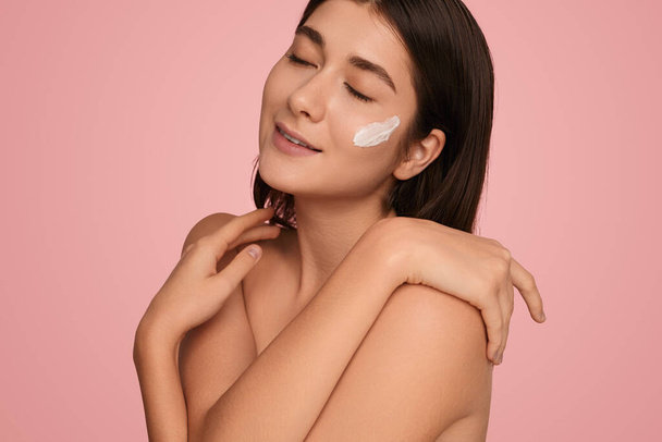 Young tender brunette with smear of moisturizing cream on cheek closing eyes and touching shoulders on pink background - Photo, Image