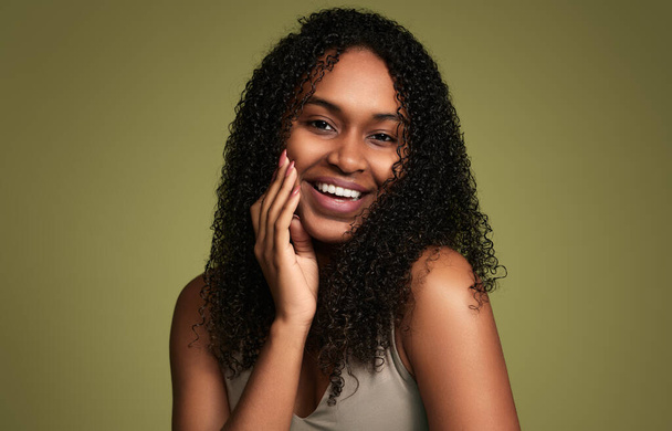 Young sensual African American female with curly hair touching soft skin of cheek and looking at camera on green background - Fotó, kép