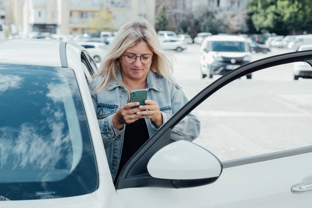 Vrouw in de buurt van auto en kijken naar haar smartphone, betalen voor parkeren en navigeren door de stad. - Foto, afbeelding