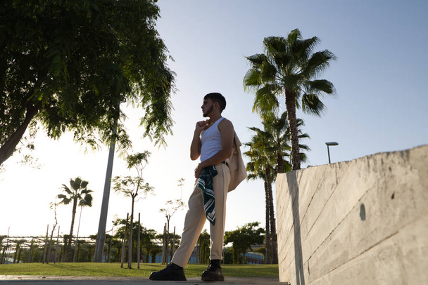 Νεαρός Λατίνος ντυμένος με μπεζ παντελόνι, λευκό t-shirt και μαύρες μπότες περπατώντας στο πάρκο και απολαμβάνοντας τη μέρα. Ο άνθρωπος έχει κοινωνικά προβλήματα και τονίζεται σκεπτόμενος τις δυσκολίες της ζωής. - Φωτογραφία, εικόνα