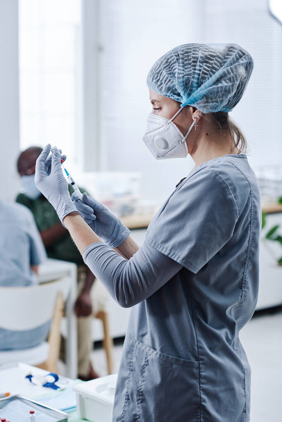 Jeune infirmière en masque et vêtements de protection préparant une seringue avec un médicament pour la vaccination debout à l'hôpital - Photo, image