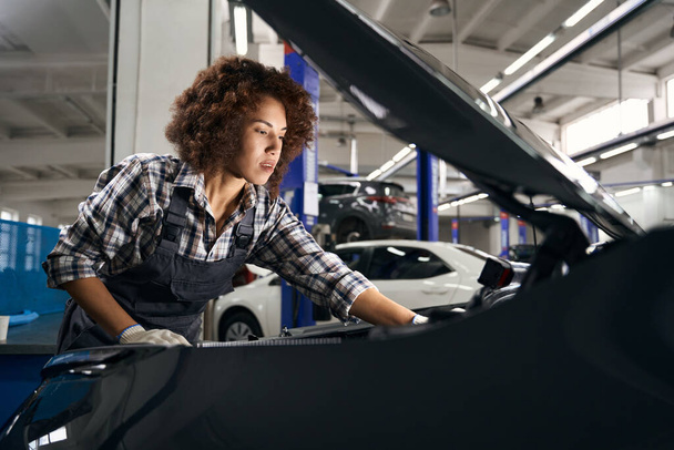 Multiraciale vrouwelijke automonteur inspecteert een auto onder de motorkap, de auto wordt onderhouden - Foto, afbeelding