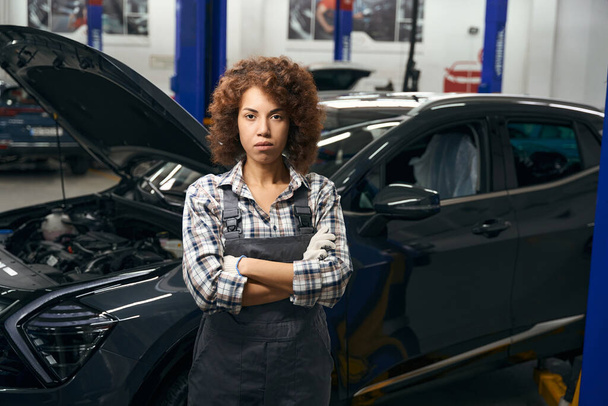 Multiracial curly employee of car repair shop stands by a car with a raised hood, the car is being serviced - Fotoğraf, Görsel
