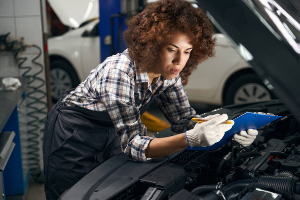 Female technician in work clothes stands near a car with an open hood, she fixes breakdowns in a blue folder - Foto, afbeelding