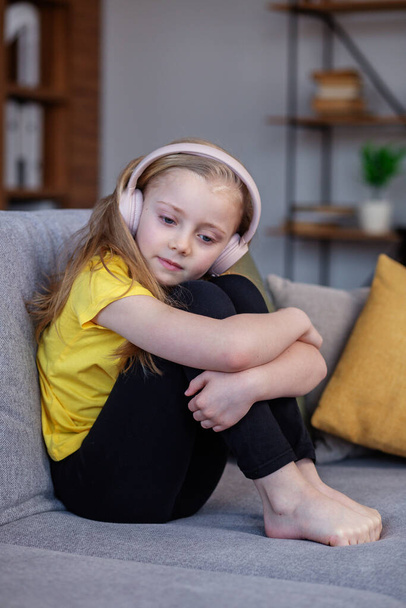 Unhappy little girl in headphones sitting in sofa at home. Concept mental health. - Foto, Imagen