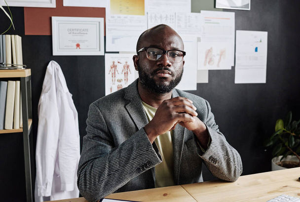 Портрет африканского врача-мужчины в очках, смотрящего в камеру, сидящего за столом и работающего в офисе - Фото, изображение