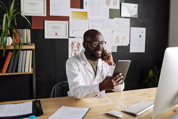 Africano médico masculino sentado en el lugar de trabajo sosteniendo la tableta PC y sonriendo mientras habla por teléfono móvil - Foto, imagen